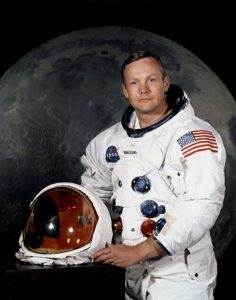 Portrait of Astronaut Neil A. Armstrong, commander of the Apollo 11 Lunar Landing mission in his space suit, with his helmet on the table in front of him. Behind him is a large photograph of the lunar surface [NASA, Public Domain; Courtesy, Wikimedia].